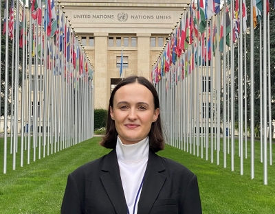 A woman with short brown hair in a black jacket and white top standing in front of the United Nations Building in Geneva