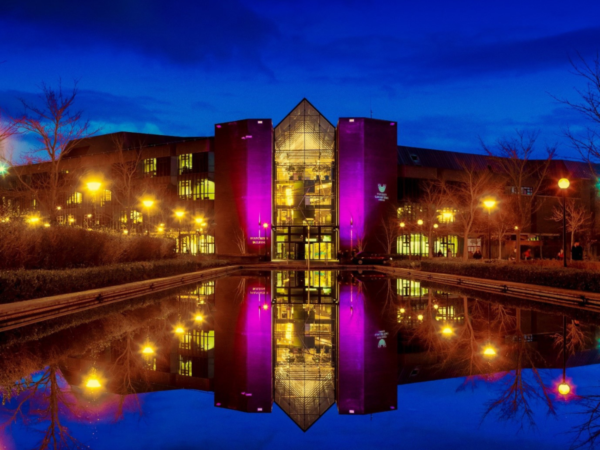 The exterior of the Foundation Building at University of Limerick, home to the University Concert Hall, lit up at night, with its image reflected in the fountain outside the Foundation Building