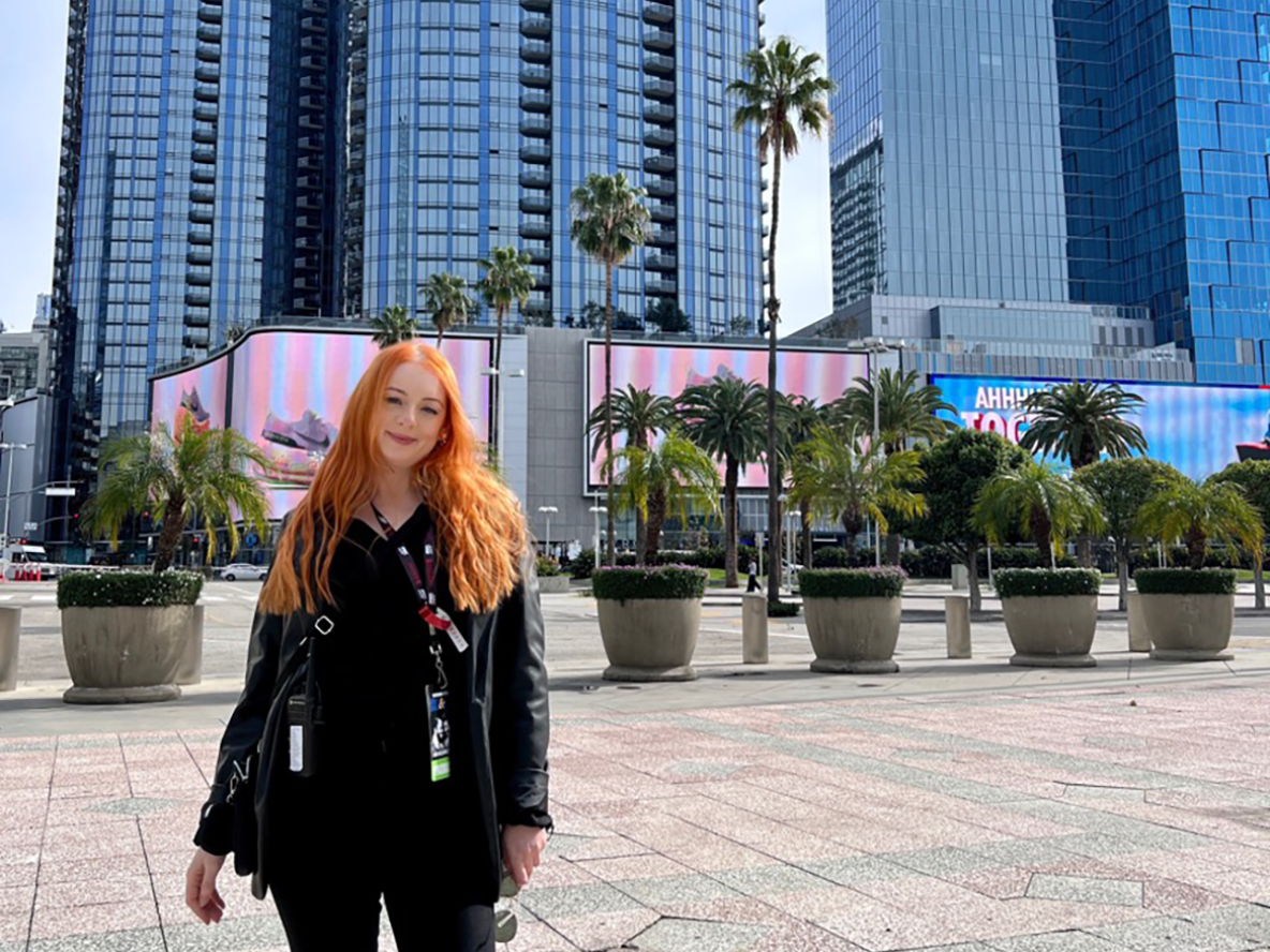 A woman with long red hair standing in front of a tall building with trees behind her