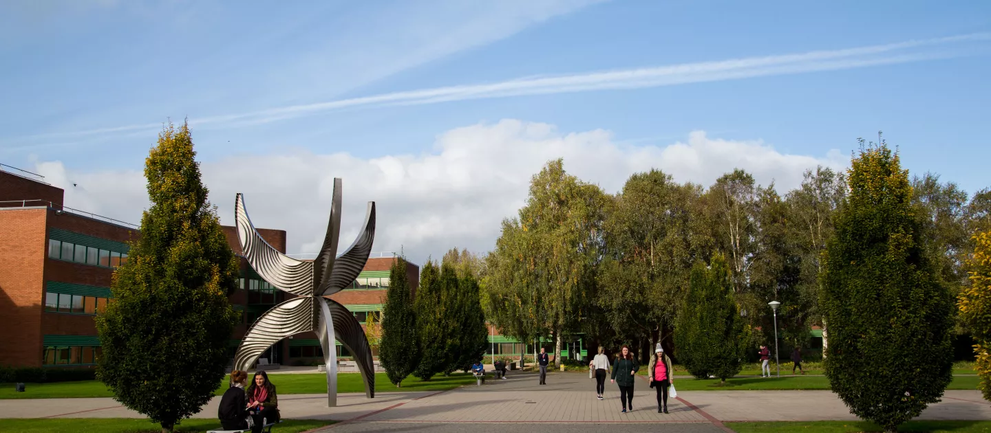 View from KBS of statue outside Schumann Building with people walking in front and trees in the background. 