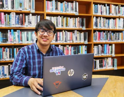 Picture of a student with a laptop at a desk, in the background is a bookshelf with a lot of books on it