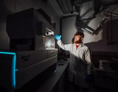 A man holds up his arm placing a petri dish into a machine in a laboratory. He is dressed in a white lab coat and protective glasses. 