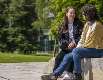 Two girls are sitting outside on a stone bench facing each other chatting. It is a sunny day and there are large trees in the background.