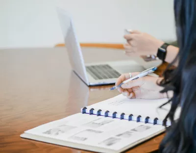 person writing in notebook with another person in background using a laptop