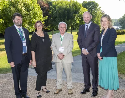 Photos of five individuals standing together with Green lawn and bushes in the background. 