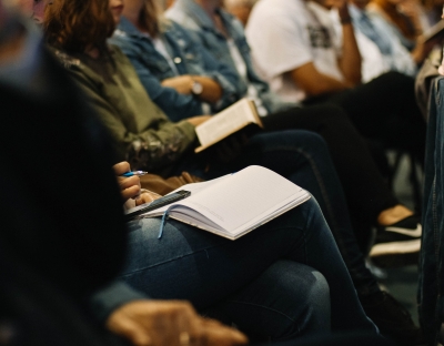 Row of people attending a conferences. They are seated and some rest notebooks on their knees.