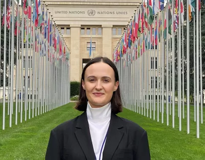 A woman with short brown hair in a black jacket and white top standing in front of the United Nations Building in Geneva