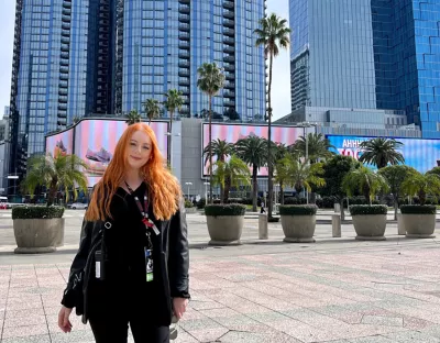 A woman with long red hair standing in front of a tall building with trees behind her