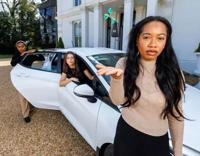 A woman standing in front of a white car throwing three green dice in the air while two women stand in the background, one sitting in the car and the other leaning on the back door