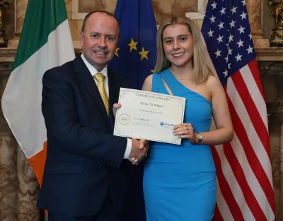 A photo of a man in a black suit, white shirt and gold tie, standing beside a women in a blue dress. They are shaking hands and holding an award certificate between them, in front of the flags of Ireland, the European Union, and the United States. Paul is wearing a black suit and gold tie, hol