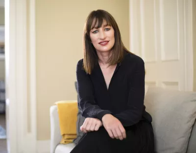 A photo of a woman wearing a black dress, sitting on a cream sofa, hands folded, against the background of a cream wall and white door.