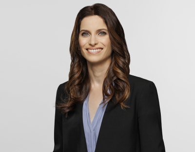 A corporate headshot of a woman with wavy brown hair wearing a black suit jacket 