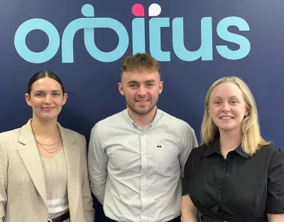 Two women and a man standing side by side in front of a wall with a sign that reads Orbitus