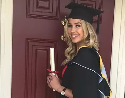 A woman with blonde hair wearing a black graduation cap and gown holding a diploma in front of a red door