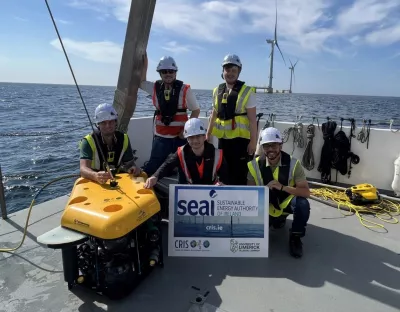 Five people wearing hard hats and hi visibility jackets and life jackets aboard a platform on an off-shore wind farm in the Atlantic Ocean with wind turbines in the background