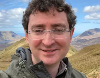 A photo of a man with glasses, wearing a jacket, smiling to the camera against a backdrop of mountains