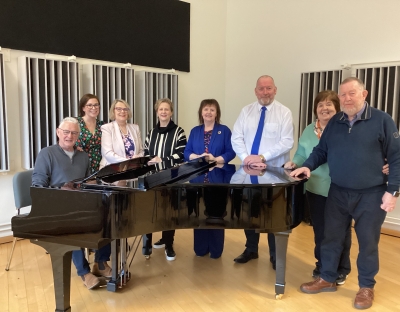 A photog of a group of eight men and women standing around a black piano