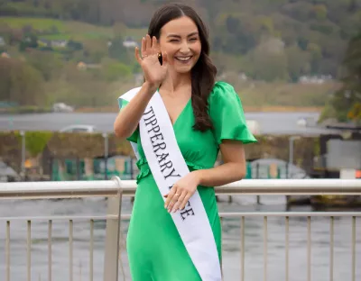 A photo of a person in a green dress and white sash, waving, with trees and a bridge over water in the background 