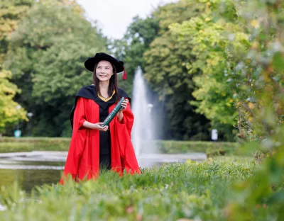 A photo of a woman wearing red graduation robes and black hat, smiling and holding a green scroll. She is standing against a backdrop of a water fountain and green trees and foliage.