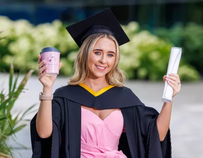 A woman in a pink dress, and black graduation cap and gown holding a diploma and pink coffee cup