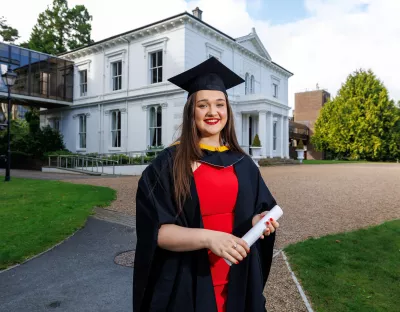 A woman in a red dress wearing a black graduation cap and gown standing in front of a white building.