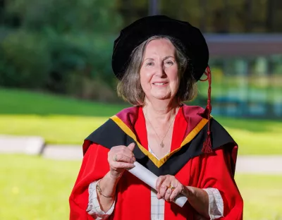 A photo of a woman in red ceremonial robes and a black graduation cap, holding a white scroll and smiling. In the background are grey steps and green grass and trees.