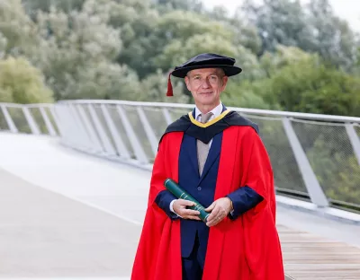 A man in a red graduation gown wearing a navy suit standing on a bridge with trees in the background