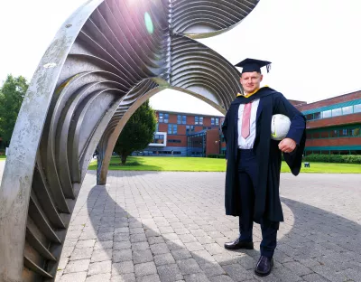 A photo of a man in a black suit, white shirt and pink tie underneath black graduation robes, wearing a graduation cap and dark shoes. He is holding a soccer ball under his left arm, standing in front of a tall metal sculpture. In the background is a red brick building with windows.