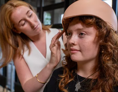 A photograph of two women with red hair. On the right, closest to the camera, is a woman in a black top sat under a pale pink helmet. She is being attended to by a woman in a white top stood behind her, who is tucking her hair into the helmet. The helmet is scalp-cooling device for people undergoing chemotherapy treatment