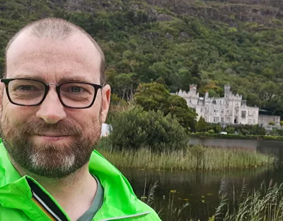 Man wearing glasses with bright green jacket on stands facing camera with an old building behind him in a lakeside location