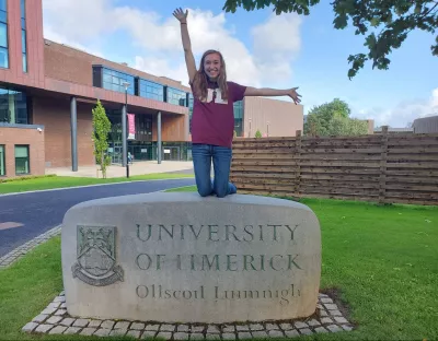 A woman in a wine t-shirt with UL on it kneeling on a University of Limerick sculpture