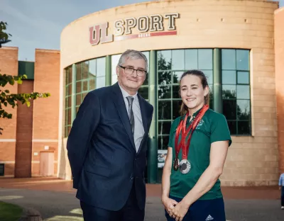 Man with glasses and wearing suit to the left and young girl to the right wearing paralmpic medals around her neck in university sports setting