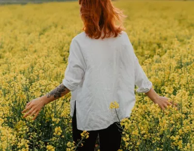 ginger haired girl with tattoo sleeve on left arm in white shirt and black pants walking through field of yellow flowers