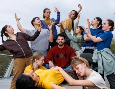 A photo of a man in a red jumper looking directly ahead, surrounded by dancers pulling various dance shapes with their arms. In the background is a bridge and green foliage.