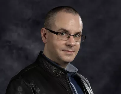 A man wearing glasses and a black leather jacket standing in front of a dark grey backdrop looking at the camera