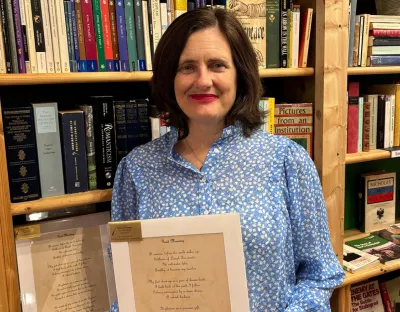 A woman with shoulder length brown hair wearing a floral white and blue shirt standing in front of a bookcase holding a printed poem.