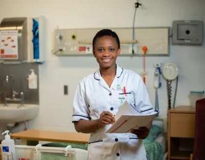 Woman wearing a white nurses uniform standing in a hospital ward holding a pen and chart