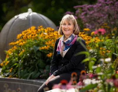 Woman with shoulder length brown hair, wearing a a black jacket and trousers and multi-coloured scarf sitting amongst outside on a sunny day surrounded by flowers and plants