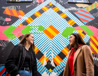 Two girls looking at each other on front of a colourful grafittied wall