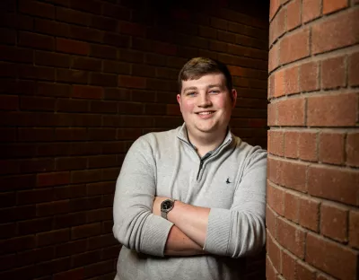 Young man leaning against a red brick wall with arms folded wearing a grey jumper