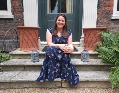 A photo of a woman with long, brown hair wearing a short-sleeved navy floral dress, sitting on a stone step. Behind her is a dark-coloured door and a terracotta pot on either side.