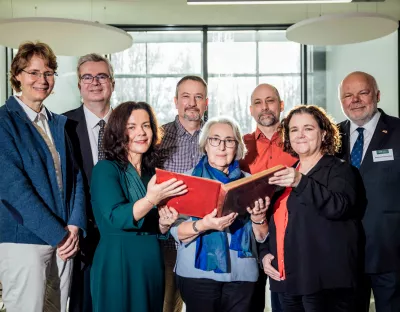 A photo of a group of people - four women and four men - standing in two rows. The three women in the front row are holding up a red book between them