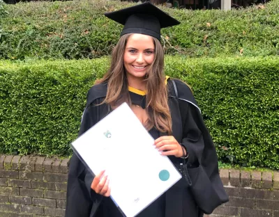 A photo of a female graduate. She is wearing black ceremonial conferring robes, holding her parchment, against a brick wall and green hedge.