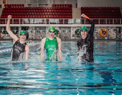 Three male triathletes wearing traithlon suits, goggles and swim hats jumping up in a large swimming pool