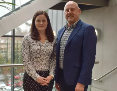 A photo of a woman and a man in smart dress, standing together against a grey wall and window.