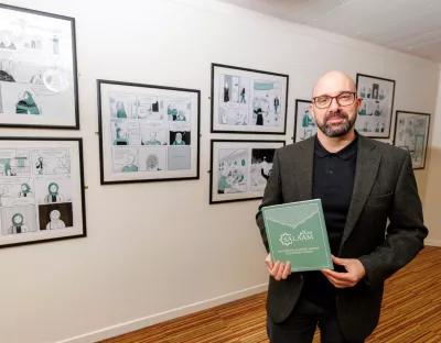 A photo of a man in smart dress, standing in front of a wall of framed pictures, holding a paper booklet.