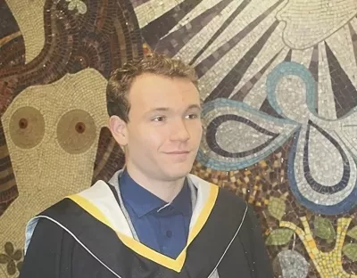 A man with blonde, short hair wearing a blue shirt and black and yellow graduation gown, standing in front of a mosaic mural looking off to the side of the camera.