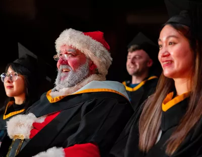 A woman with dark hair and glasses, a man dressed in a Santa suit, and a woman with long brown hair, all sitting in a row looking to the side of the camera wearing graduation gowns. 