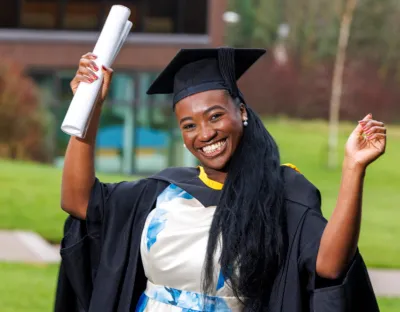 A photo of a women smiling, with her hands in the air, wearing black ceremonial robes and cap over a blue and white dress. She is holding a white scroll in one hand.