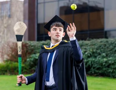 A photo of a man dressed in dark conferring robes and cap, holding a hurley and throwing a sliotar with the other hand